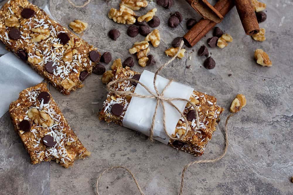 Chewy date granola bars topped with chocolate, chopped walnuts, and shredded coconut, surrounded by scattered walnuts and chocolate chips