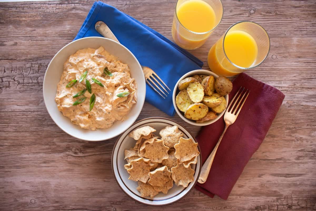 a bowl of buffalo chicken dip with tortilla chips, potatoes and two glasses of orange juice. 

