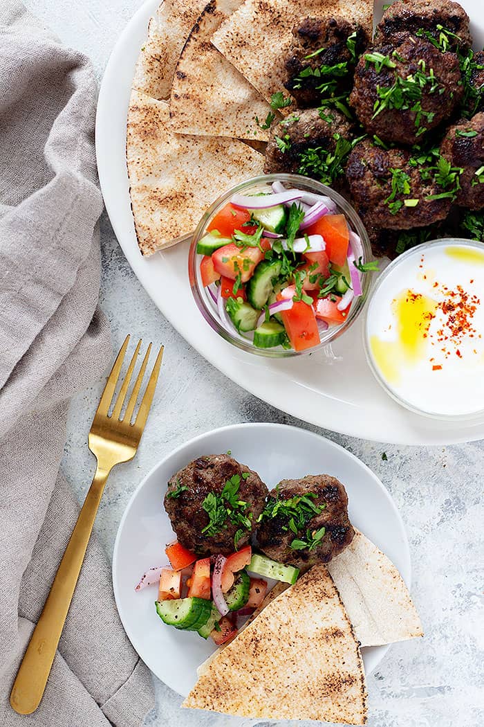 Tow plates of kofte kabobs with cucumbers tomatoes and yogurt sauce and pita bread. 
