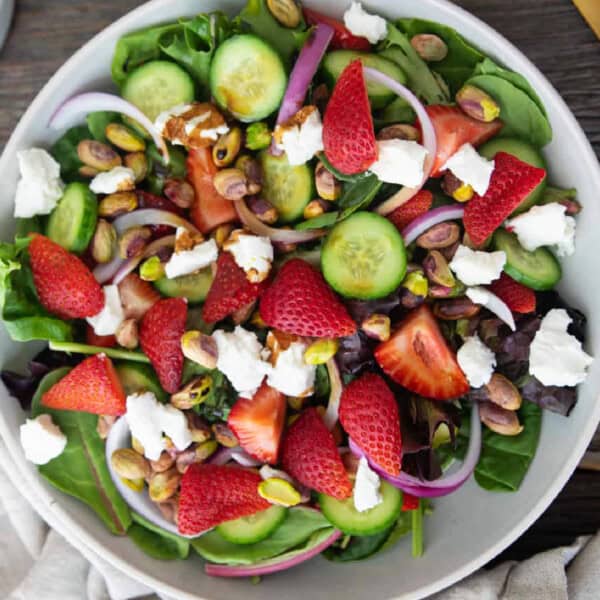 A vibrant strawberry goat cheese salad in a large white bowl, featuring fresh sliced strawberries, creamy crumbles of goat cheese, crunchy toasted pecans, and crisp mixed greens, all drizzled with a glossy balsamic glaze.