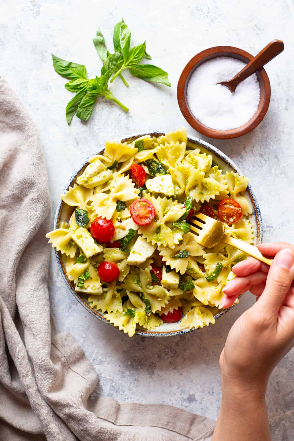 A bowl of pasta salad on a white background with salt and basil. 