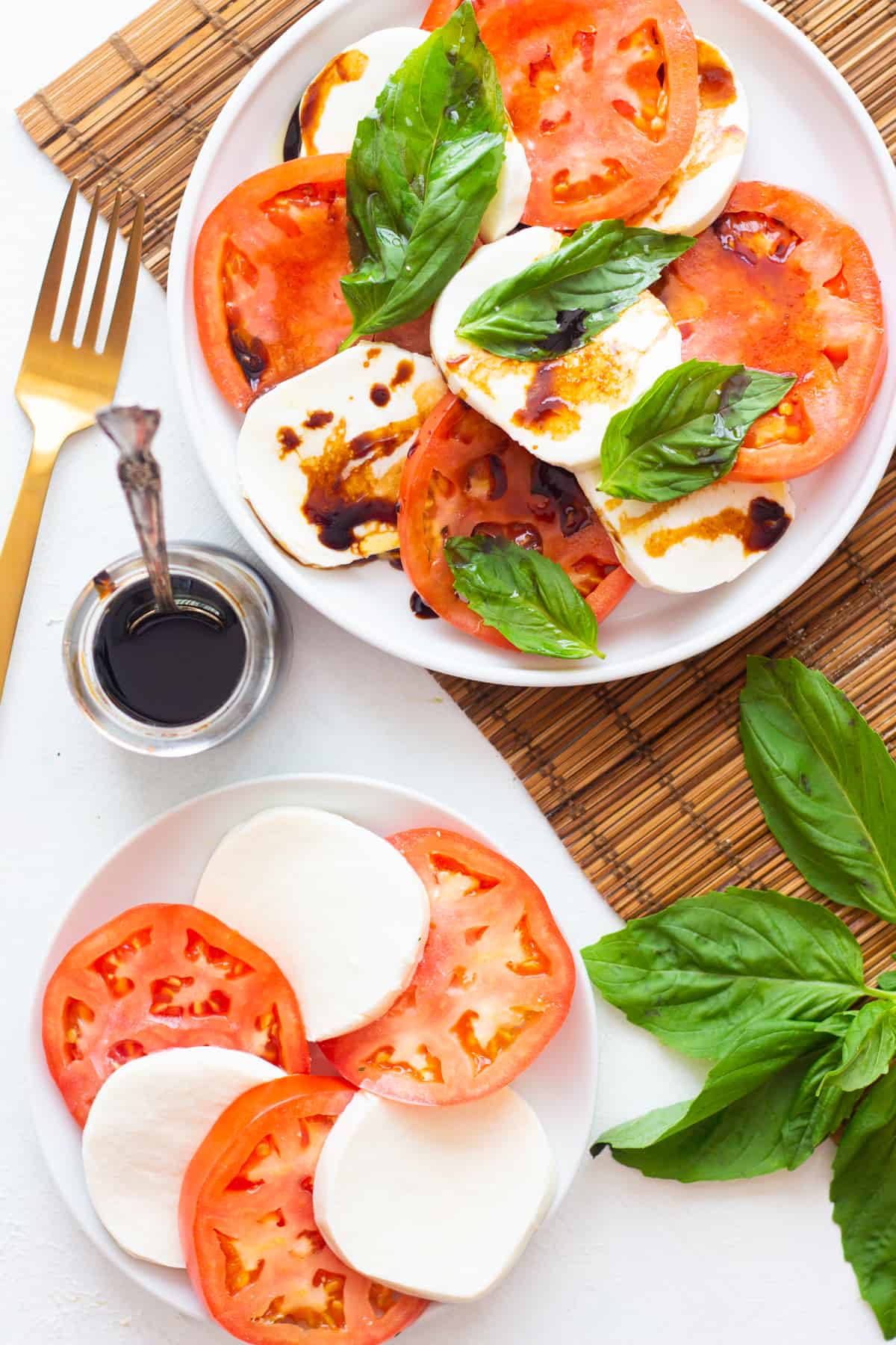 Two plates on a white surface, one has tomatoes and mozzarella and the other has caprese salad topped with balsamic vinegar. 