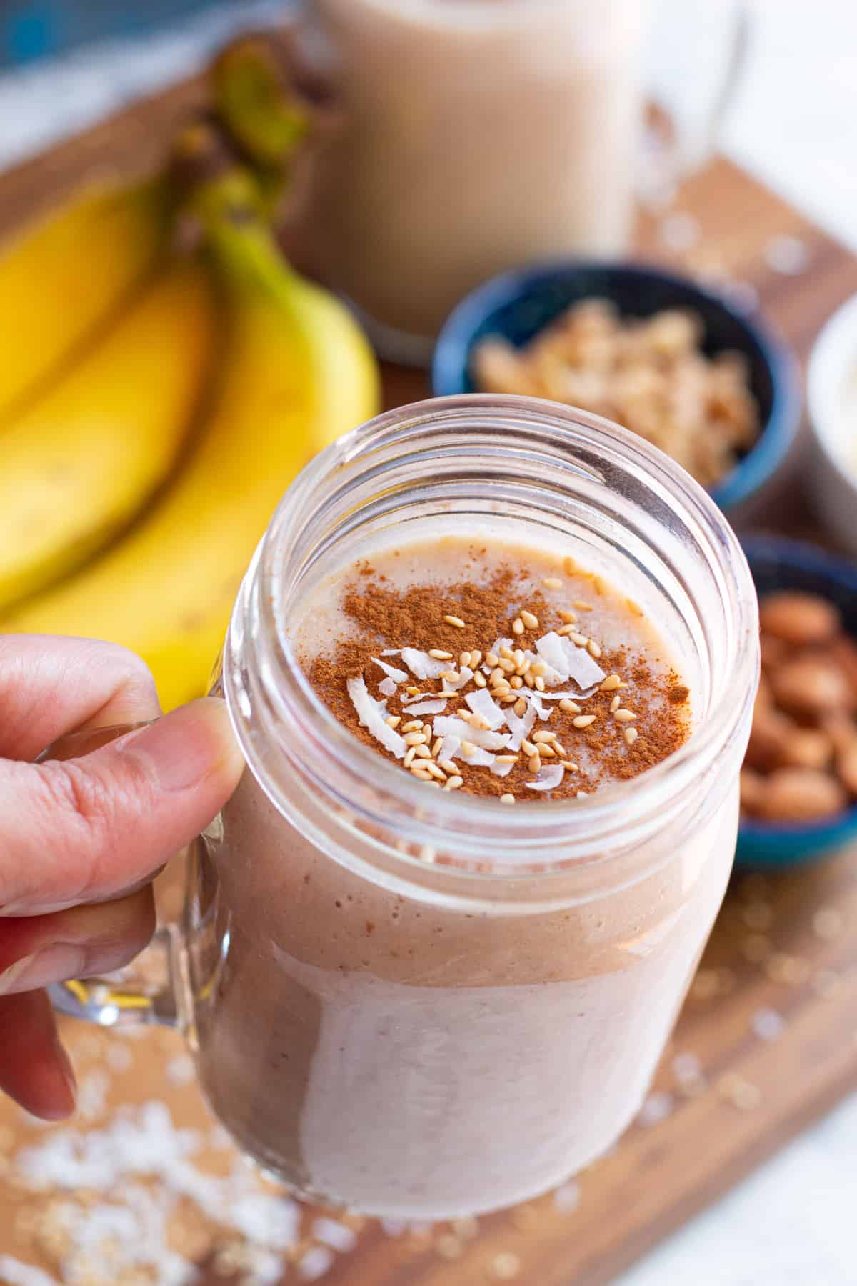 A hand holding a glass of date banana shake. In the background we have bananas and nuts.