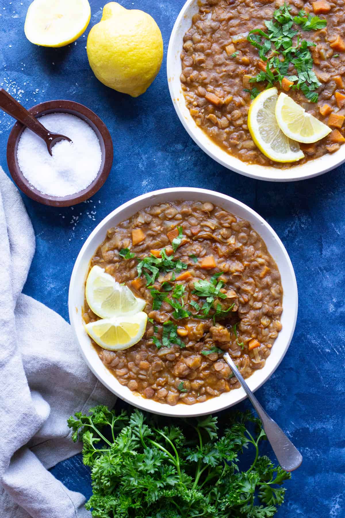 two bowl of lentil vegetable soup with parsley and lemon and salt on a blue board.