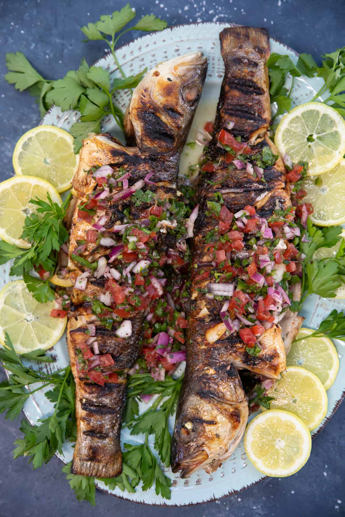 overhead shot of two grilled branzinos on a platter with lemon and herb garnish. 