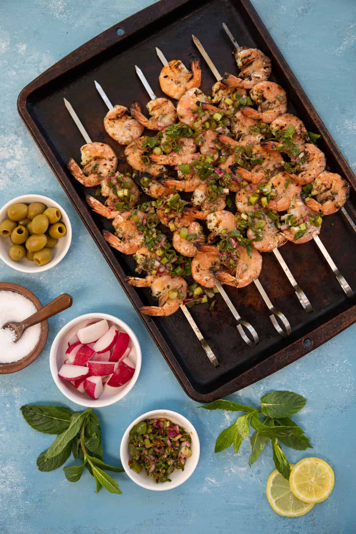 Grilled shrimp skewers on a baking sheet on a blue backdrop. 