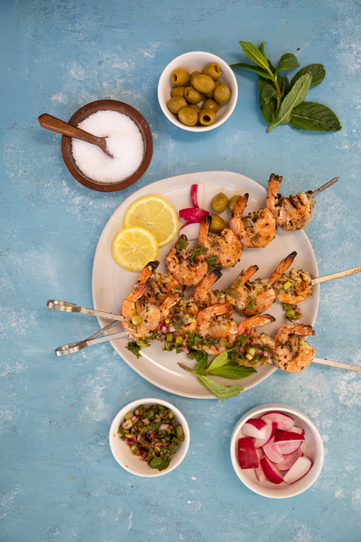 a round plate with prawns on metal skewers. Three bowls with olives, radishes and sauce.