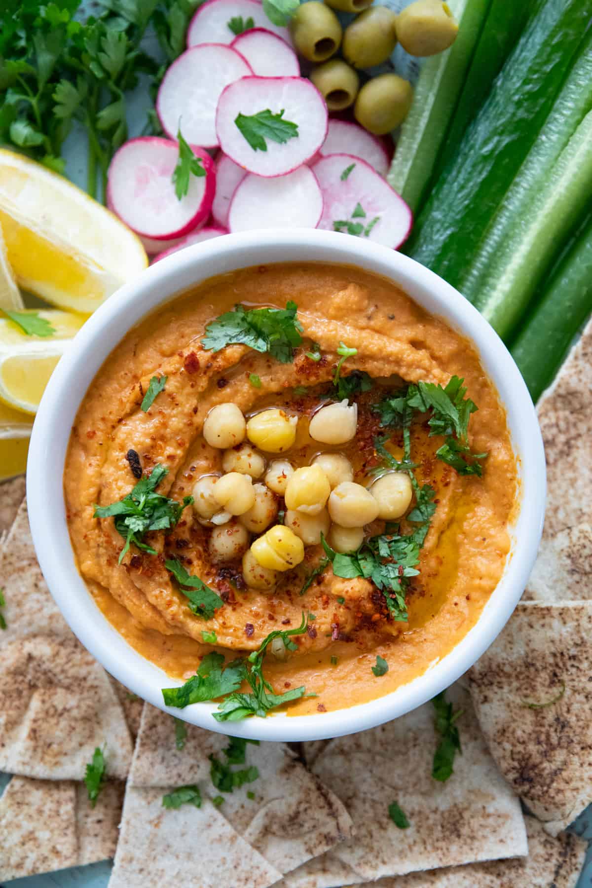 bowl of roasted red pepper hummus topped with chickpeas and olive oil and herbs on a board with pita and vegetables.