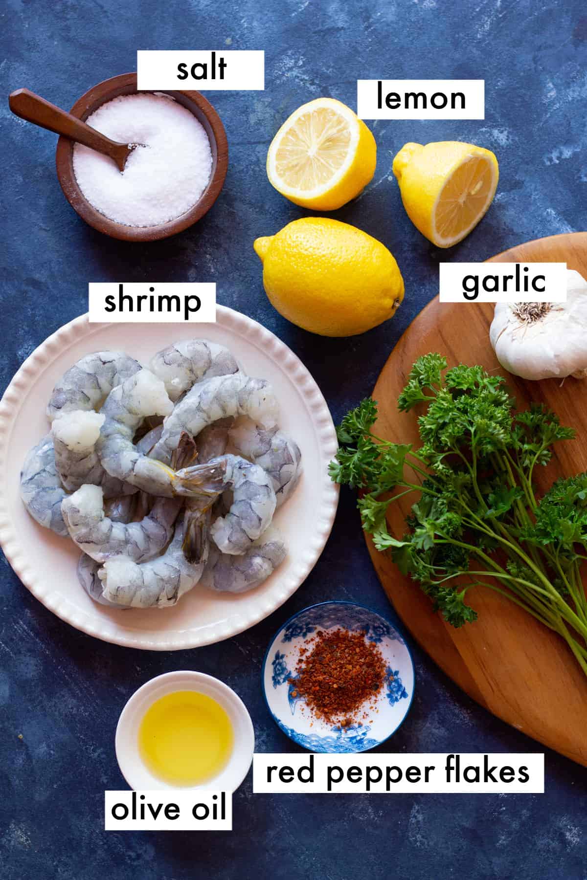 shrimp, olive oil, garlic, red pepper flakes, salt and lemon on a dark blue backdrop.