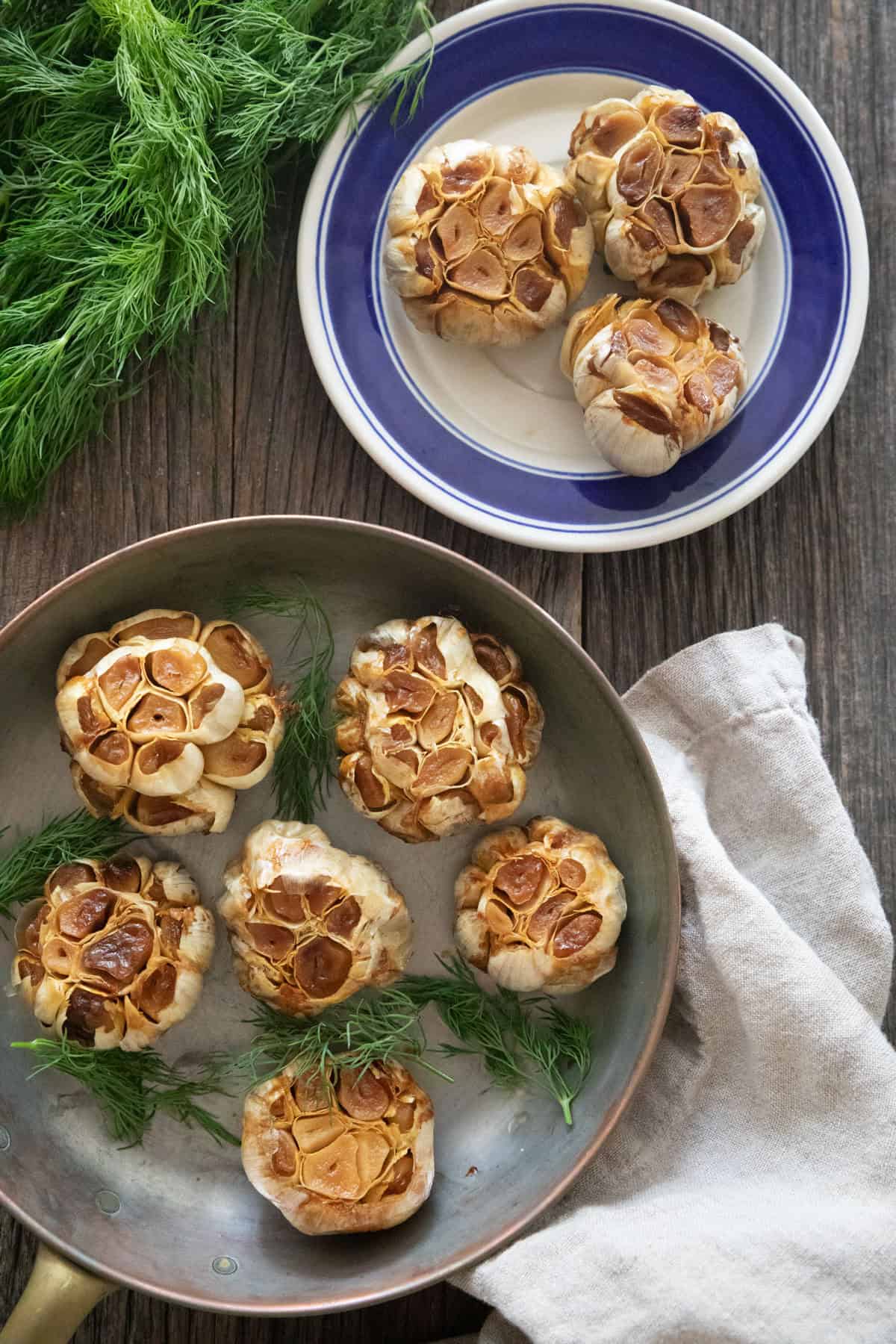 roasted garlic on a plate and in a pan on a wooden backdrop.