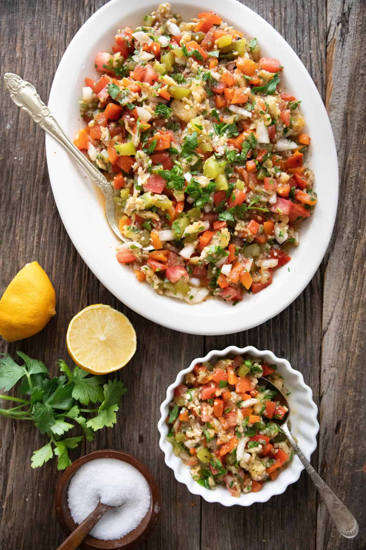 A platter of smoky eggplant salad with a bowl on a wooden backdrop with lemon and herbs. 