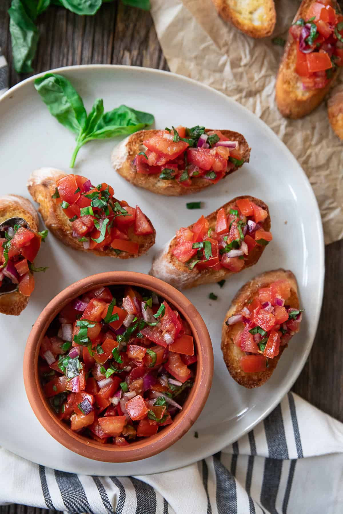 Authentic and classic Italian Bruschetta served on a classic white plate with a small bowl of the bruschetta mix and basil as garnish