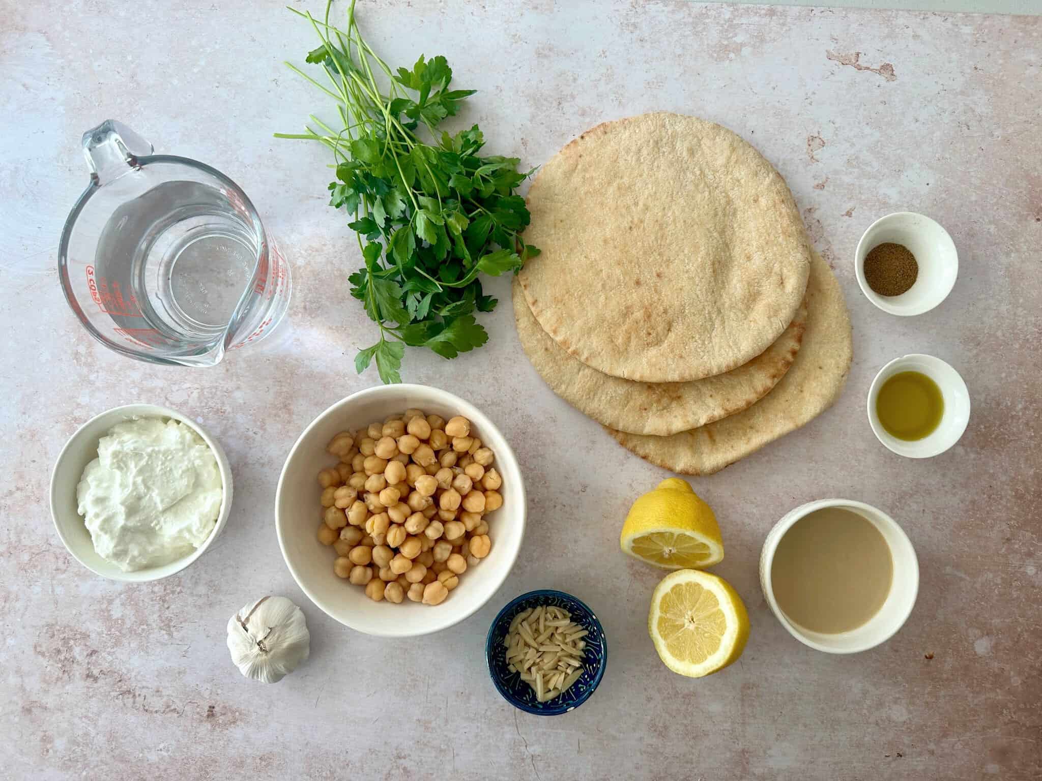 Fatteh recipe ingredients you will need: Pita, chickpeas, yogurt, garlic, fresh parsley, olive oil, tahini, lemon juice, cumin, black pepper, slithered almonds, and water