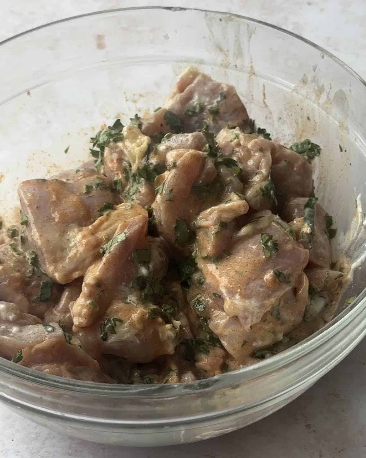 Marinated Mediterranean chicken skewers in a glass bowl. The marinade ingredients, including olive oil, yogurt, lemon juice, tomato paste, parsley, cilantro, cumin, coriander, paprika, salt, and pepper, are visible on top of the chicken cubes. 