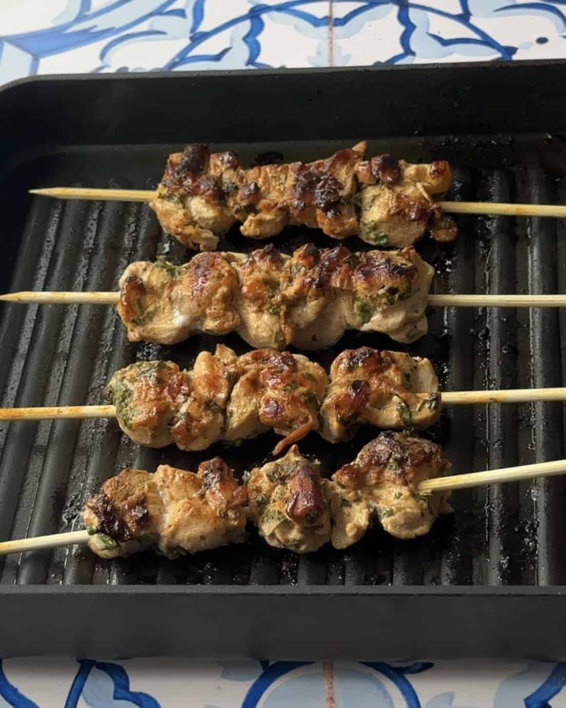 Grilled Mediterranean chicken skewers, showing their golden-brown color, cooking on the grill. The skewers are arranged in rows, with colorful char marks visible on the surface of the chicken pieces.