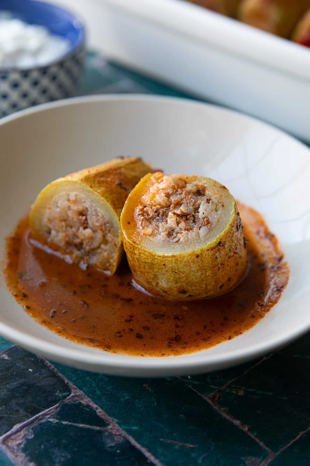 one Turkish stuffed zucchini served in a white bowl. It's sliced in the middle to show off the stuffing and is sitting in its beautiful tomato sauce