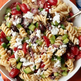 Greek pasta salad ingredients: Fusilli pasta, ripe tomatoes, cucumbers, red onions, Kalamata olives, feta cheese, Greek dressing.