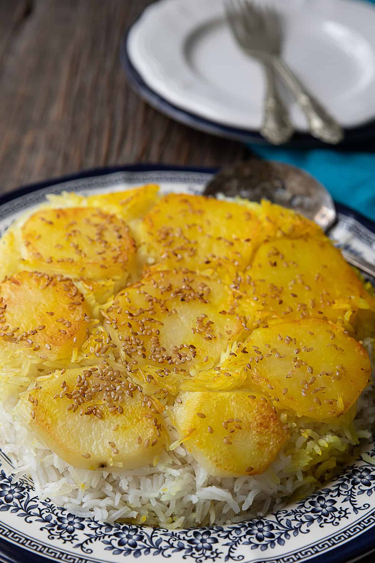 A close-up of Persian potato tahdig: Layers of thinly sliced golden potatoes forming a crispy crust over fragrant saffron-infused rice, served in a traditional Iranian pot.