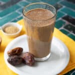 date coffee smoothie served in transparent glass cup with one on a white serving dish with 3 dates next to it and on a yellow cloth napkin with a small ramekin of brown sugar
