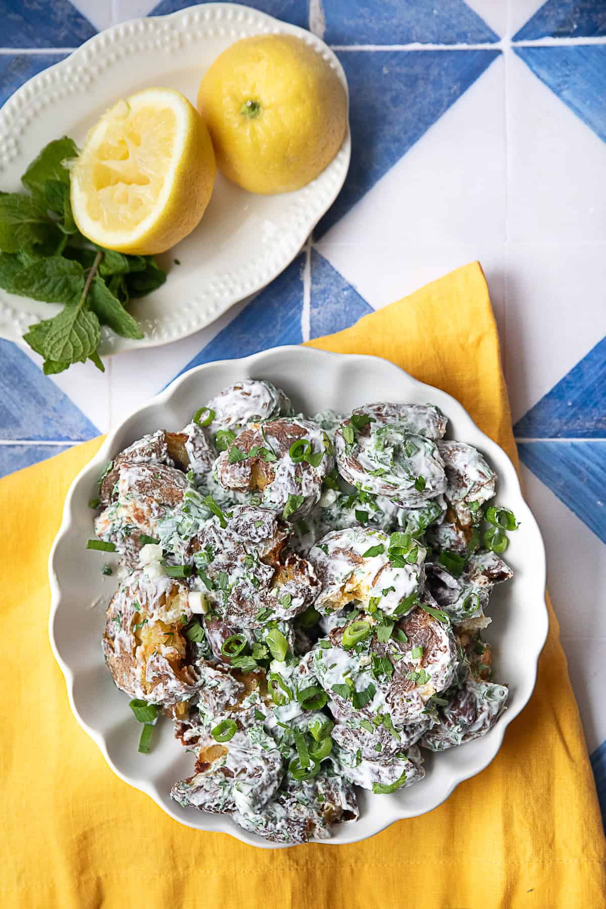 Smashed Potato Salad with Herbs Smashed potato salad served in a white bowl, featuring golden, crispy baby potatoes mixed with a tangy yogurt-tahini dressing, and topped with vibrant chopped herbs and Kalamata olives.