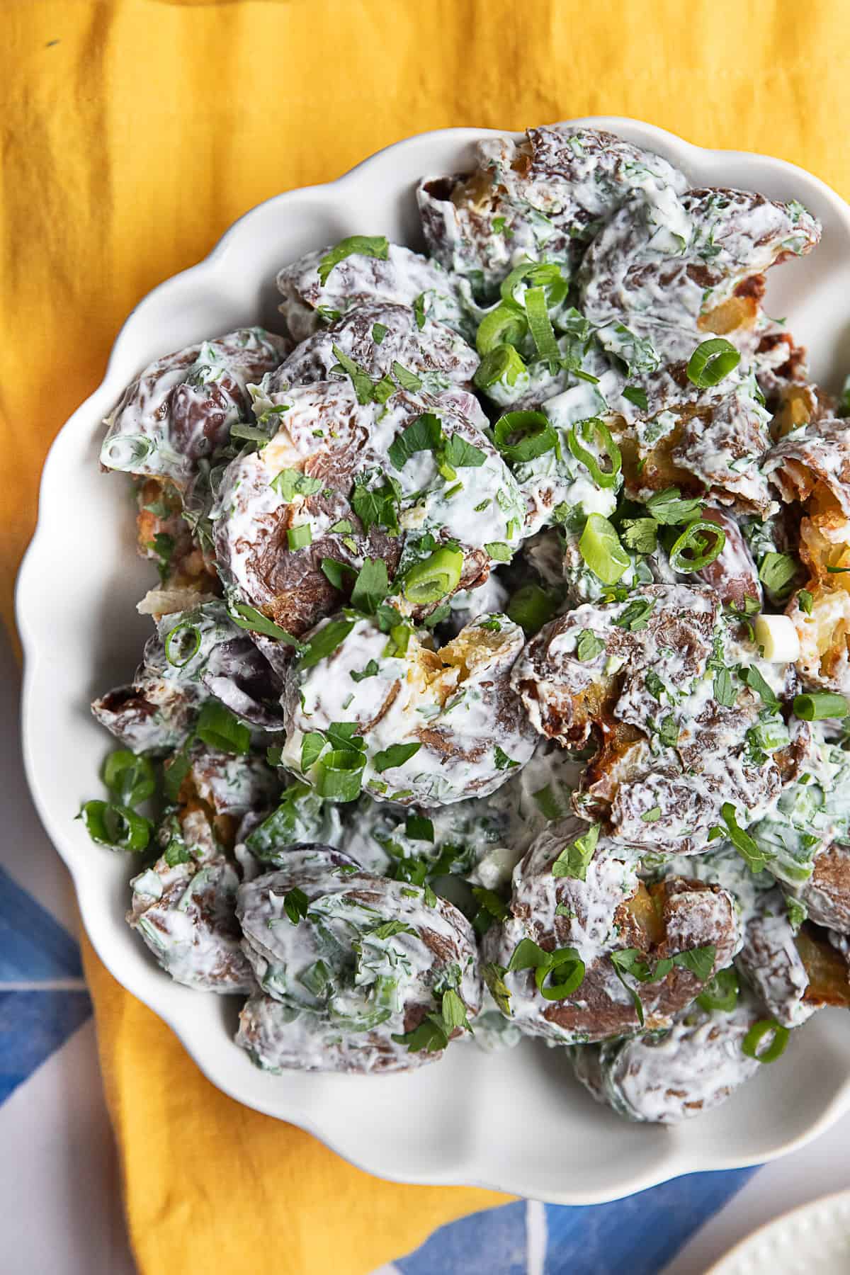 Crispy Smashed Potato Salad A white bowl brimming with smashed potato salad, featuring crispy roasted baby potatoes dressed in a creamy yogurt-tahini sauce. Freshly chopped herbs and sliced Kalamata olives add a pop of color and flavor.