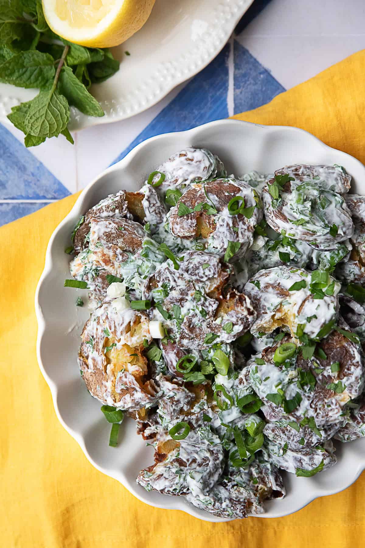 Smashed Potato Salad Overhead view of a white bowl filled with smashed potato salad. The crispy, golden-brown baby potatoes are topped with a creamy yogurt-tahini dressing and garnished with chopped mint, parsley, green onions, and sliced Kalamata olives.