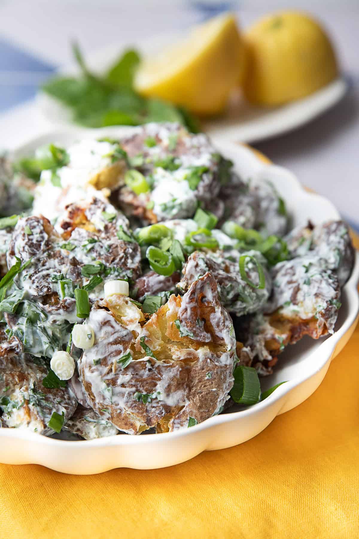 White bowl filled with smashed potato salad, highlighting the contrast between the crispy roasted potatoes and the creamy yogurt-tahini dressing, with a colorful garnish of fresh herbs and olives.