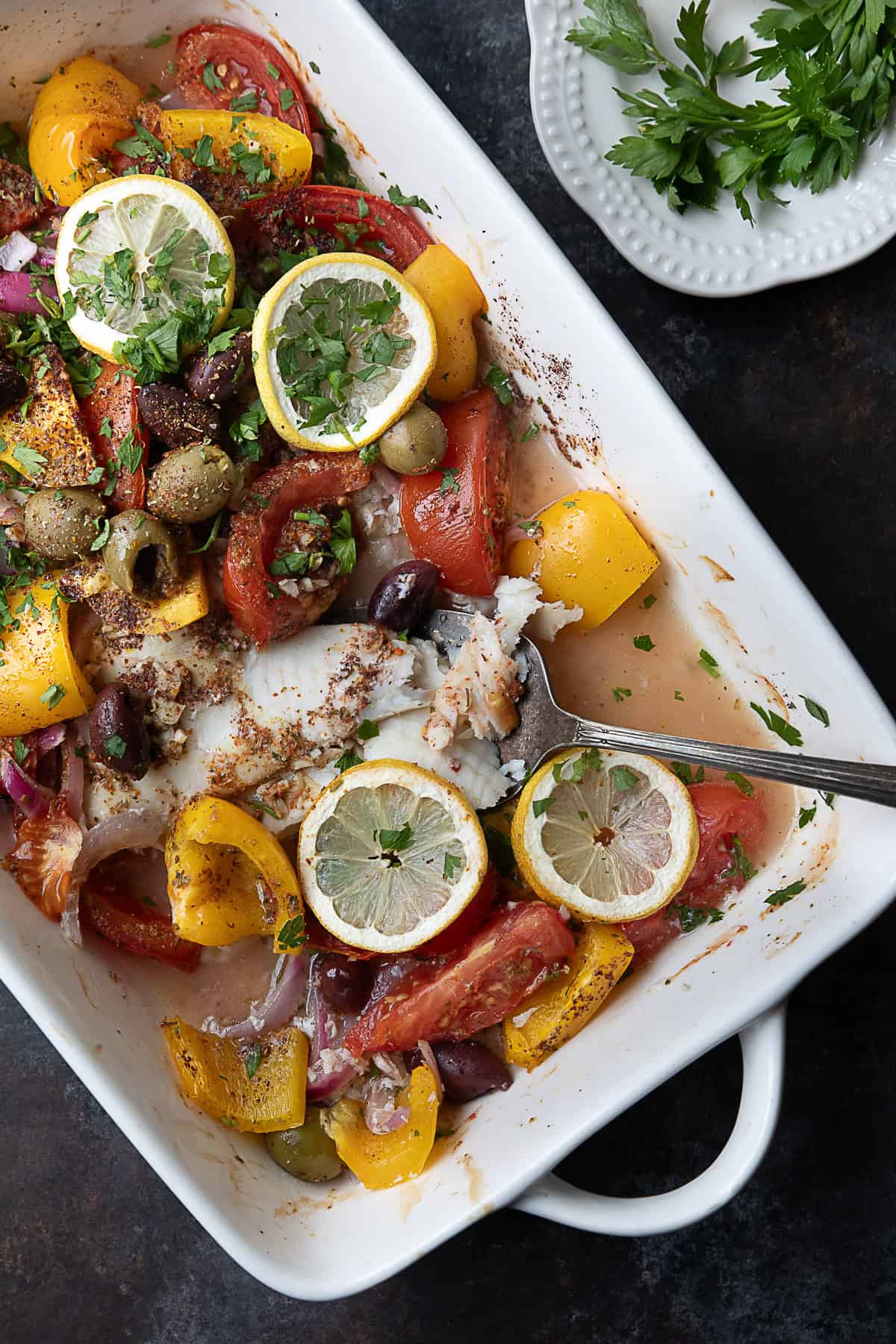Lemo Herb Flounder Baked flounder surrounded by sliced bell peppers, tomatoes, red onions, and olives, topped with lemon slices and a blend of spices in a baking dish.