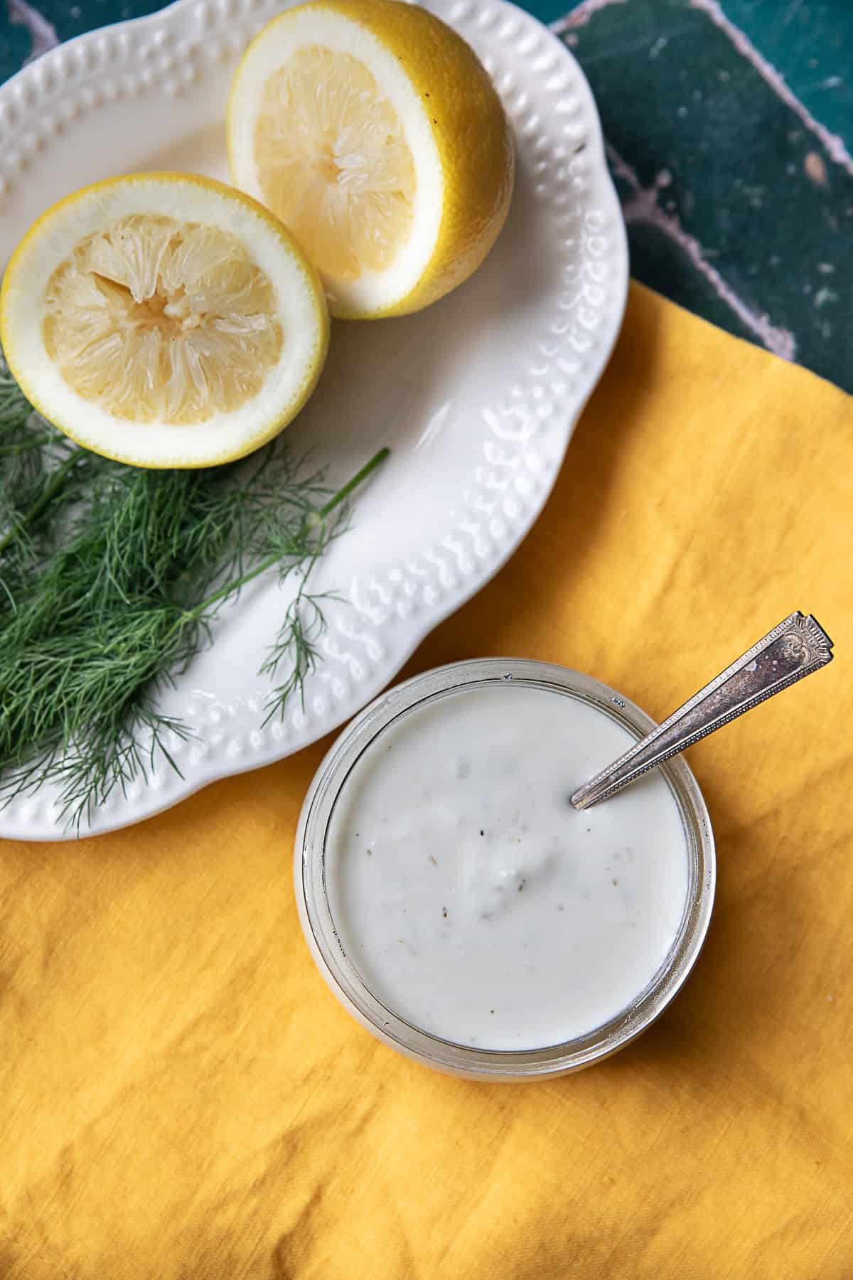 Creamy Tangy Feta Dressing a top view of a feta dressing glass jar placed next to a white plate of lemon halves and green herbs