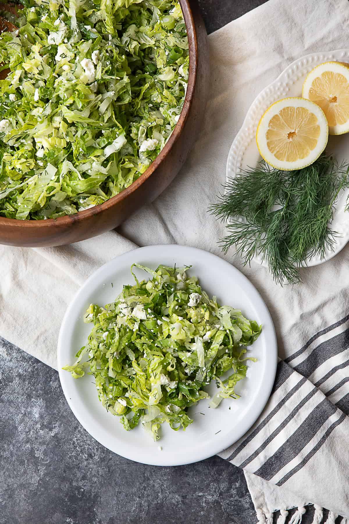 Maroulosalata Recipe A picture featuring a wooden bowl filled with Maroulosalata, a plate also filled with the salad, and another plate holding two lemon halves and some dill.