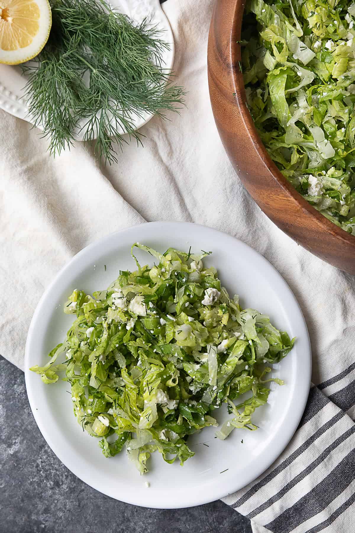 Authentic Maroulosalata A picture featuring a wooden bowl filled with Maroulosalata, a plate also filled with the salad, and another plate holding two lemon halves and some dill.