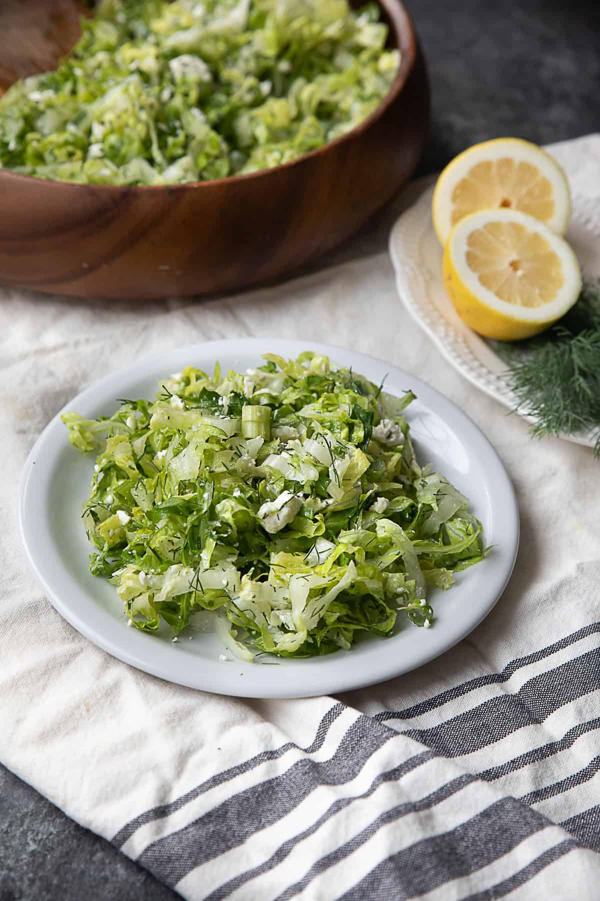 Classic Maroulosalata A picture featuring a wooden bowl filled with Maroulosalata, a plate also filled with the salad, and another plate holding two lemon halves and some dill.