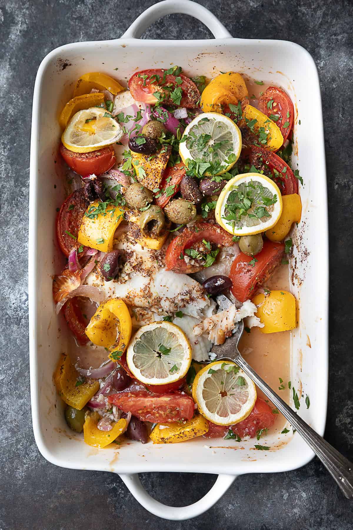Baked Flounder Baked flounder fillets layered with colorful vegetables, olives, and lemon slices, drizzled with olive oil and sprinkled with spices, served in a white oven dish.