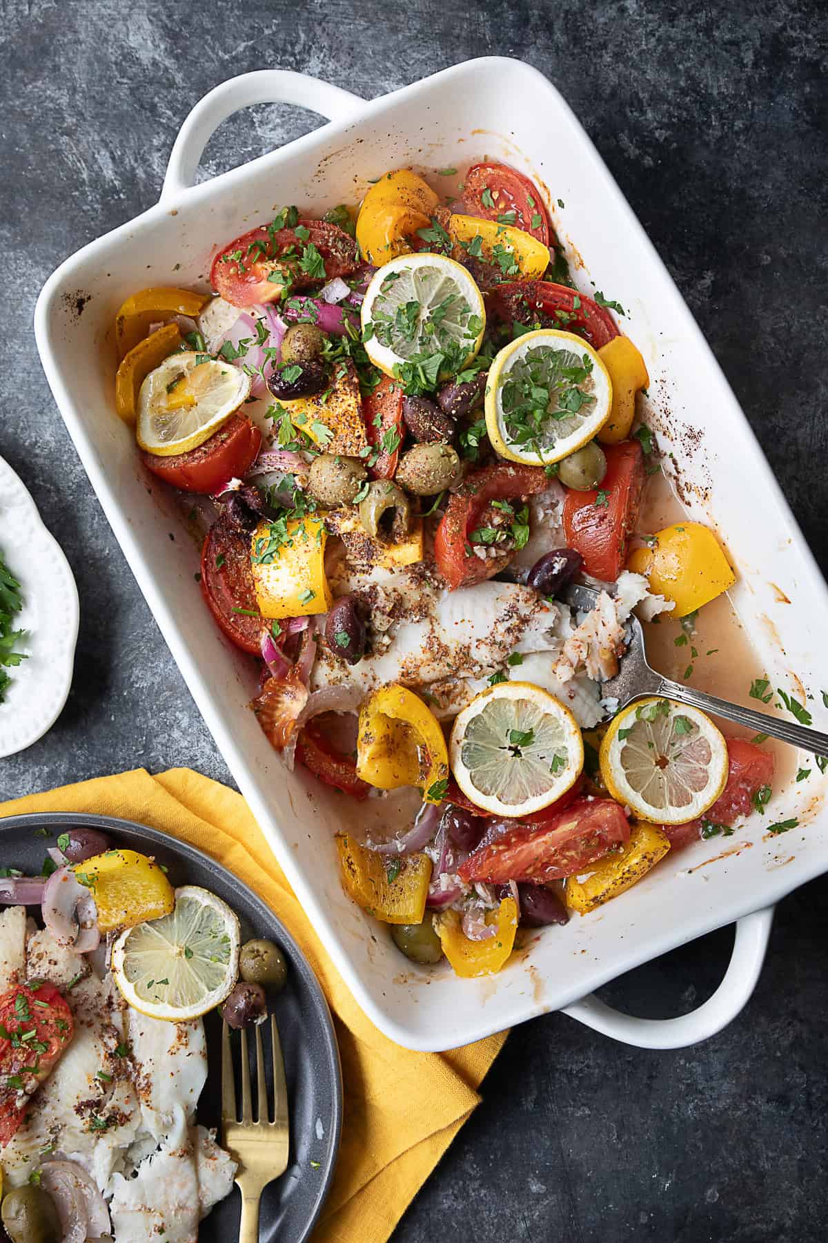 Easy Baked Flounder Baked flounder fillets topped with sliced tomatoes, bell peppers, red onions, olives, and lemon slices, garnished with herbs and spices, in a white baking dish.