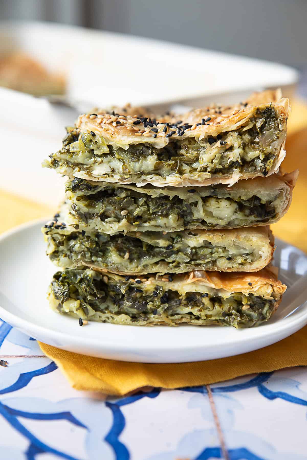 Spinach Feta Borek A stack of golden, flaky Spinach Borek slices, filled with a savory mixture of spinach and feta, topped with sesame and nigella seeds, placed on a white plate. The crispy layers of phyllo dough and the rich, green filling are visible in the close-up.
