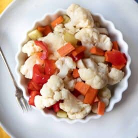 A white bowl of homemade Giardiniera with vibrant cauliflower, carrots, celery, and red bell peppers, marinated in a tangy vinegar and olive oil brine, ready to enjoy