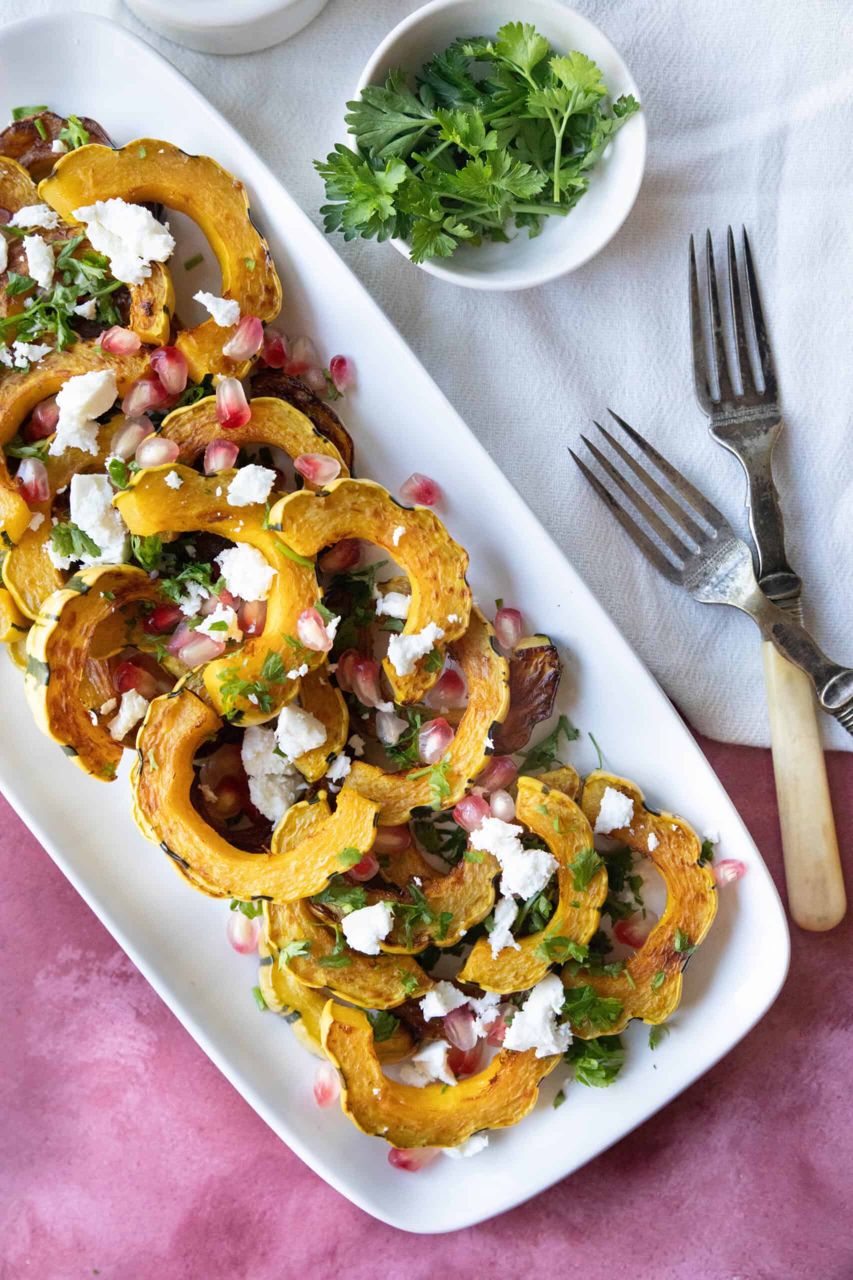 Mediterranean Roasted Delicata Squash roasted delicata squash served on a white platter with feta and pomegranate seeds sprinkled on top with some parsley as garnish