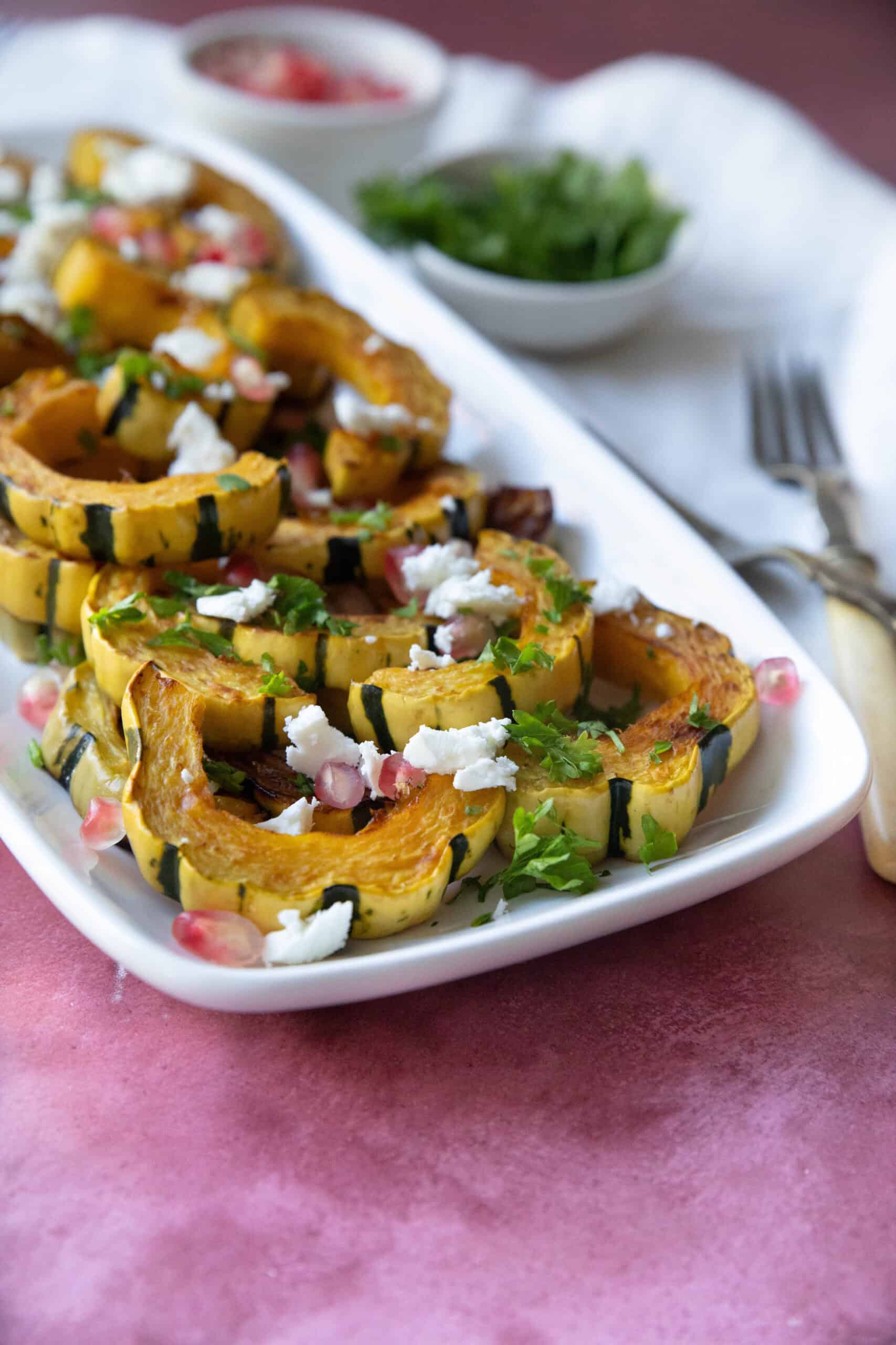 Best Roasted Delicata Squash Recipe roasted delicata squash served on a white platter with feta and pomegranate seeds sprinkled on top with some parsley as garnish