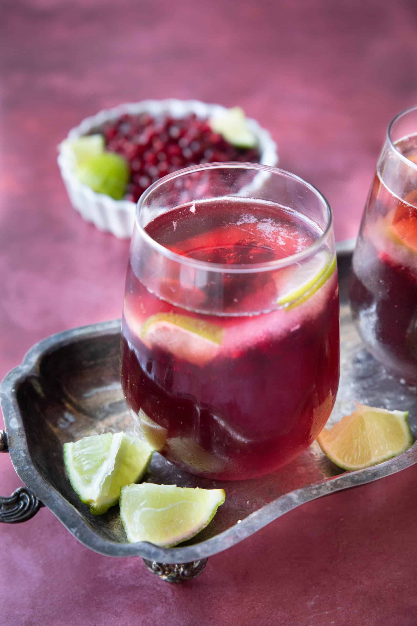 pomegranate mocktail served in a transparent glass on a silver tray with lime wedges and pomegranate arils in a white bowl in the back