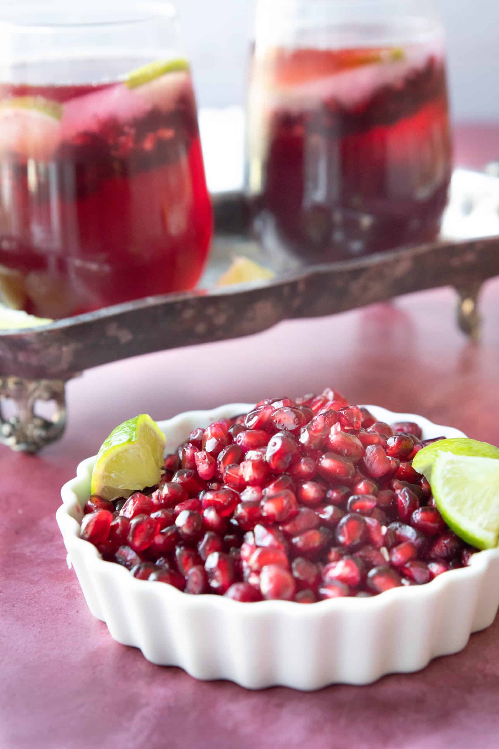 Easy Pomegranate Mocktail pomegranate arils in a small white bowl with lime wedges with two pomegranate mocktails on a silver tray