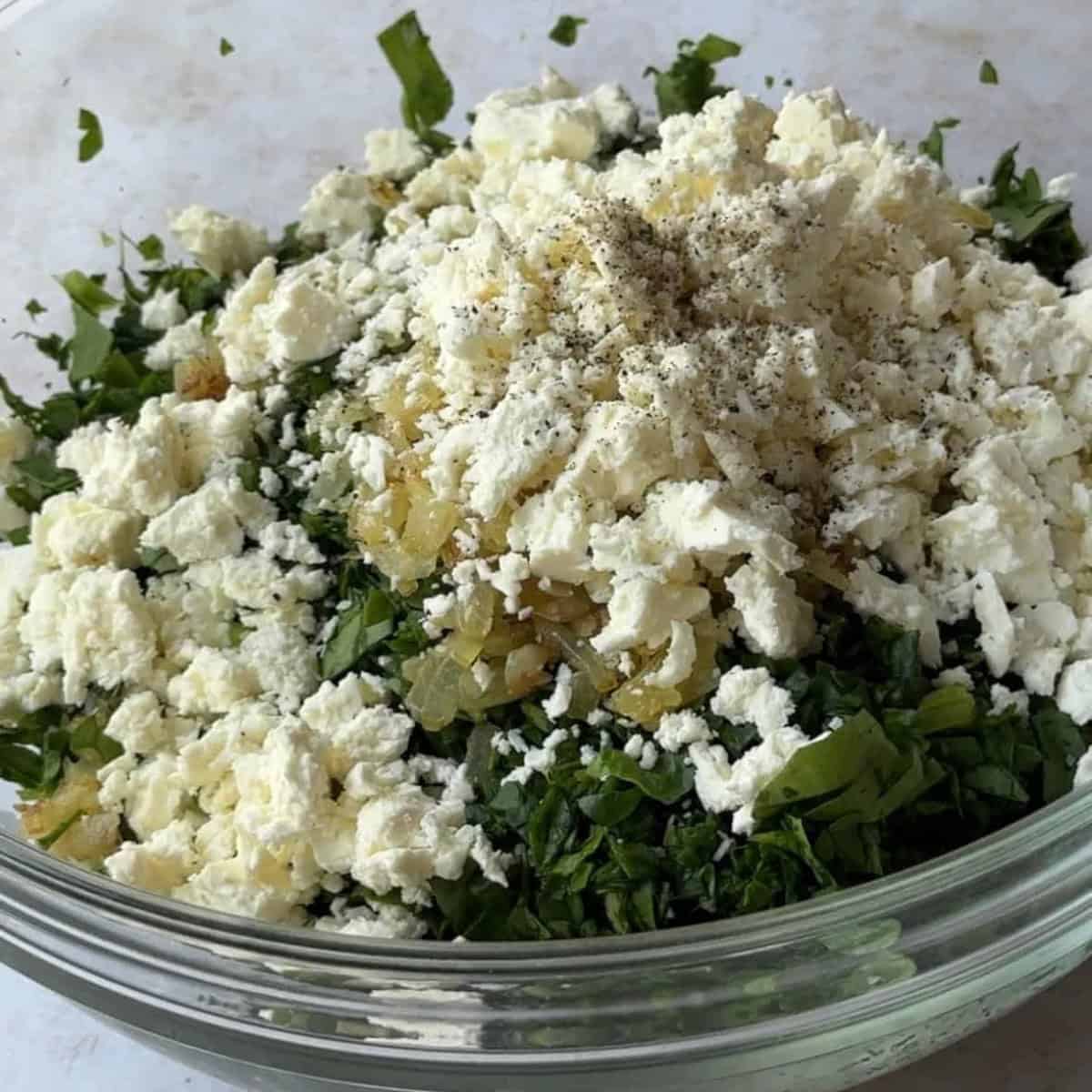 How-to-Make-Spinach-Feta-Borek A bowl filled with finely chopped spinach, sautéed diced onion, crumbled feta cheese, and a few sprinkles of salt and pepper.