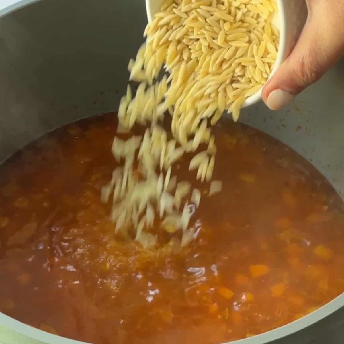 Adding uncooked orzo to the soup mixture to cook in the simmering broth.