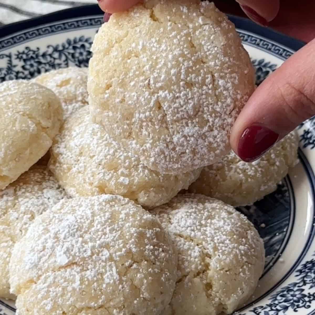 How to Make Amaretti Cookies amaretti cookies served in a decorative navy blue and white dish with one cookie held up to be seen up close