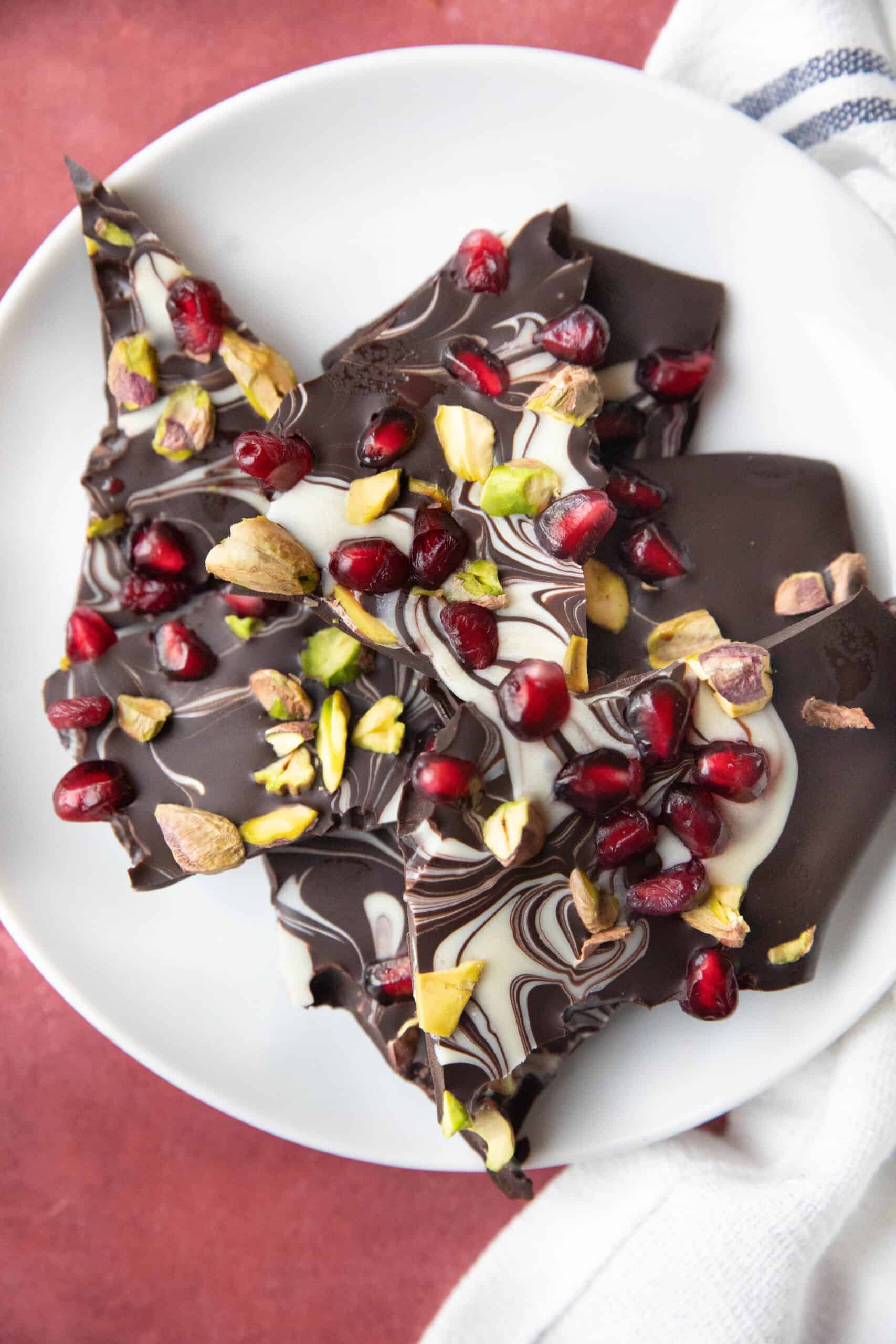 Pomegranate and Pistachios Chocolate Bark A white plate filled with large pieces of chocolate bark topped with pistachios and pomegranate seeds.