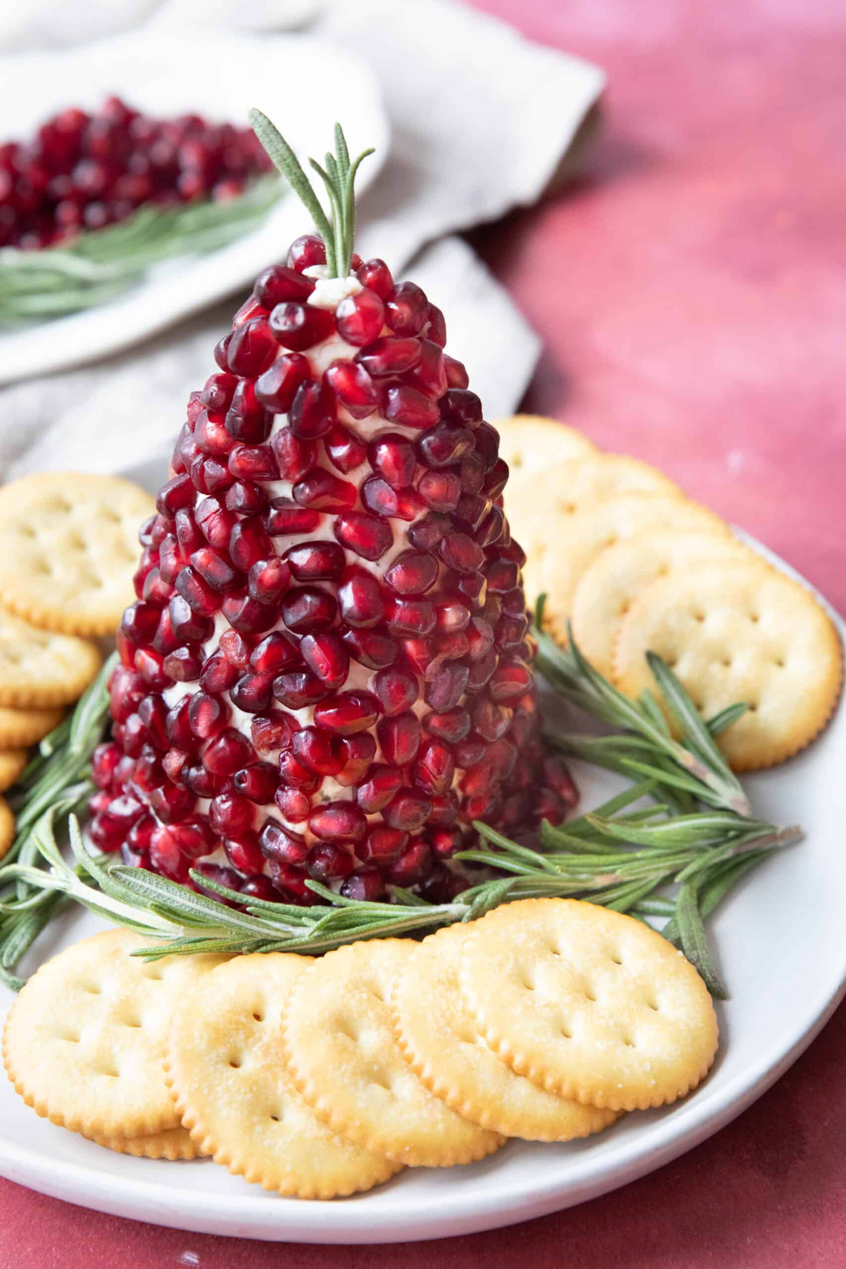 Christmas Cheese Ball Platter Christmas tree cheese ball served on a plate with sprigs of rosemary and crackers around it with a plate of pomegranate arils and rosemary in the background