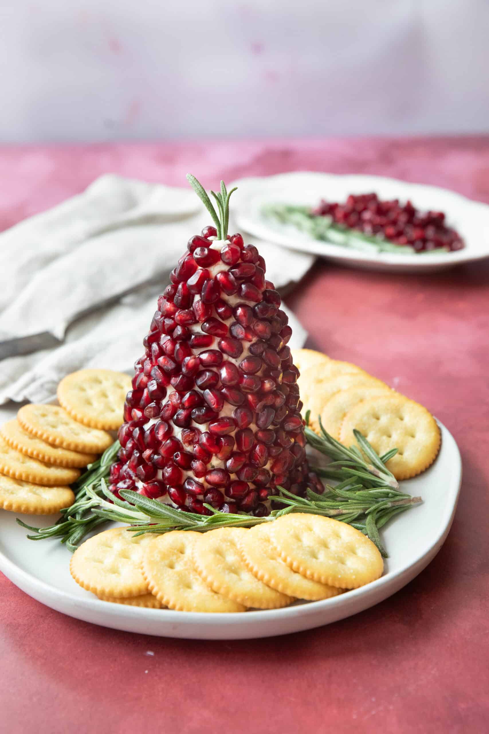 Christmas Tree Boursin Cheese Ball Christmas tree cheese ball served on a plate with sprigs of rosemary and crackers around it with a plate of pomegranate arils and rosemary in the background