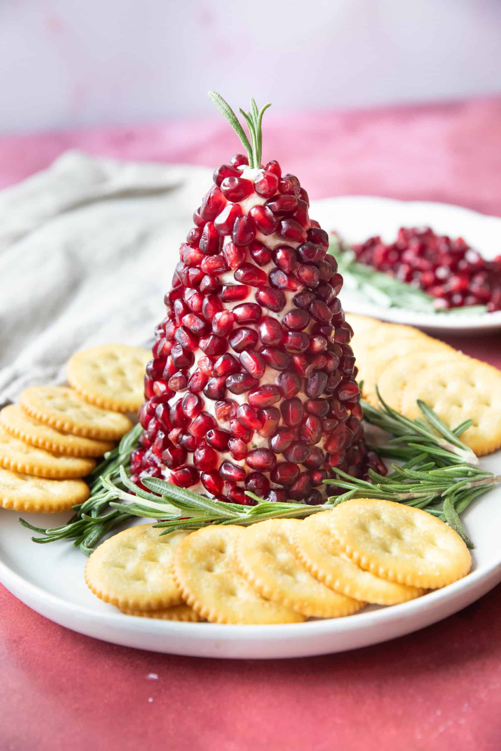 Christmas Tree Cheese Ball Christmas tree cheese ball served on a plate with sprigs of rosemary and crackers around it with a plate of pomegranate arils and rosemary in the background