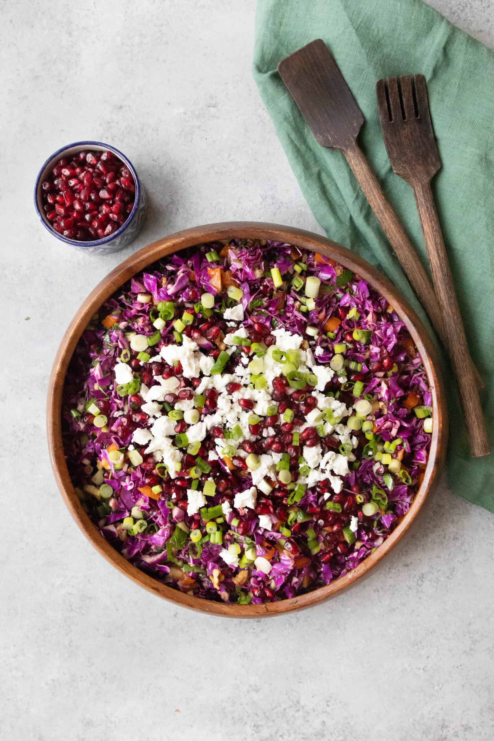 Easy Red Cabbage Pomegranate Salad red cabbage pomegranate salad served in a wooden bowl with wooden serving spoons to its right and a small bowl of pomegranate arils to its back left