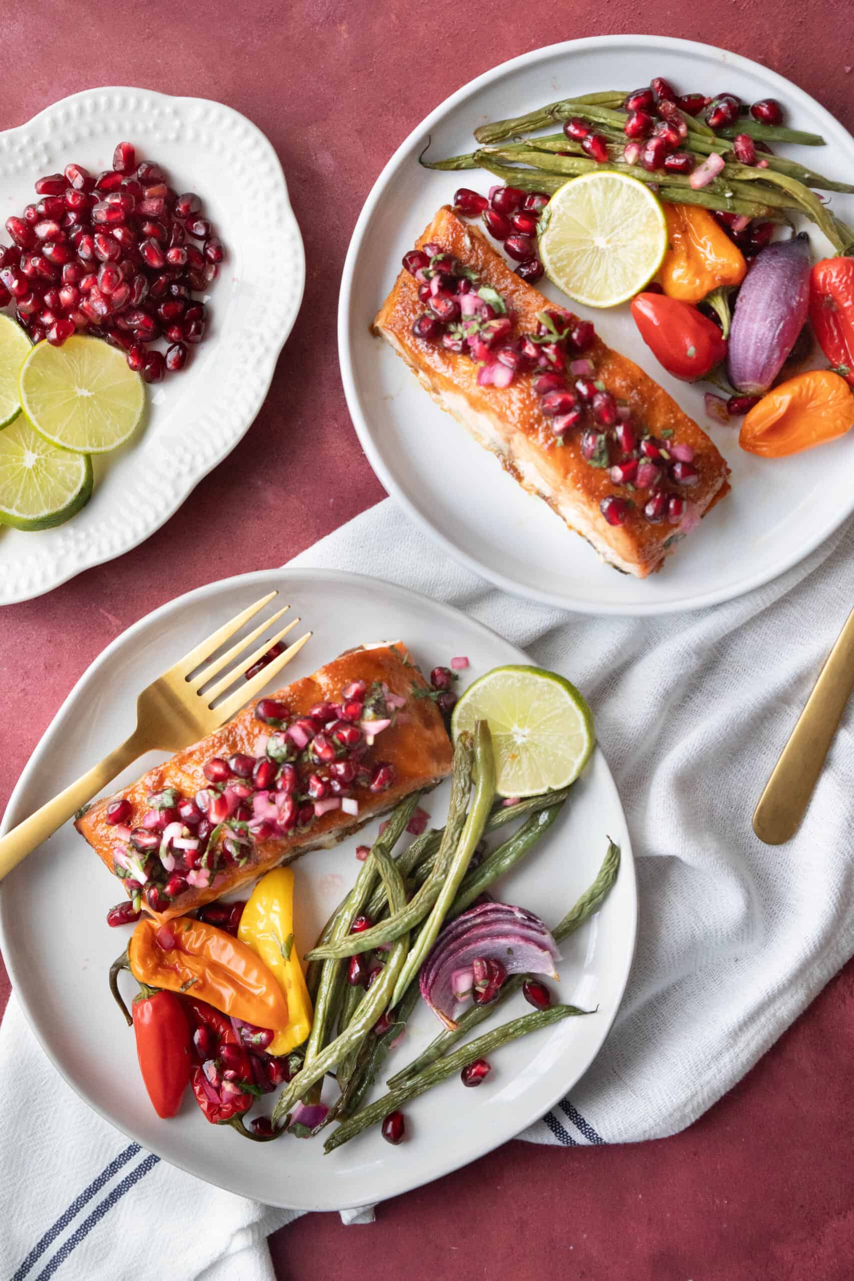 sheet pan salmon served up in two dishes with pomegranate salsa on top and beans, peppers, and onions on the slide with lime wedges and a plate of pomegranate arils and lime slices