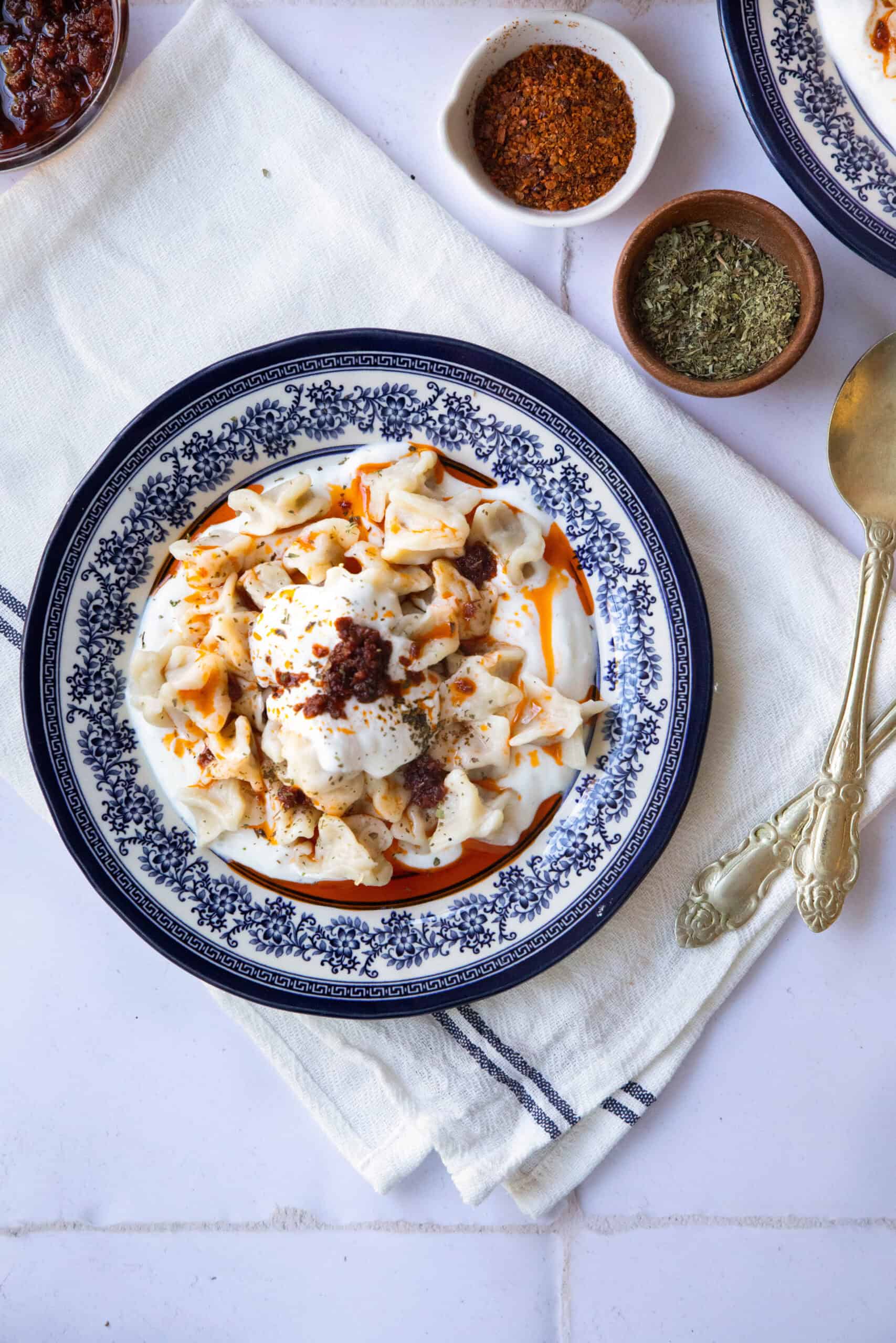 Manti A close-up view of a single serving of manti dumplings, highlighting the golden yogurt topping, vibrant red sauce, and soft dough encasing the spiced beef filling, served with dried mint and Aleppo pepper as garnish. there are also two small bowls of dried mint and cayenne pepper